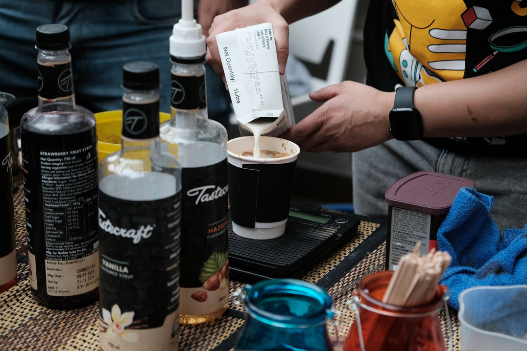 Barista pouring milk into a coffee drink beside flavored syrup bottles
